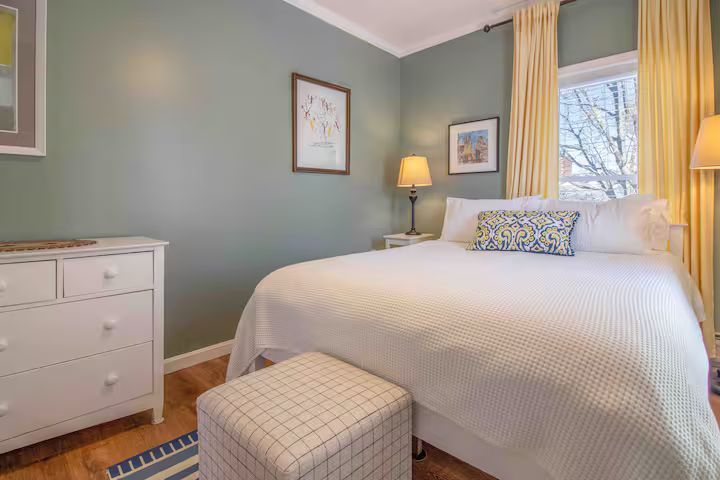 Cozy bedroom with a white bed, mint walls, dresser, lamp, and patterned ottoman by the window.