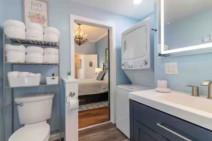 Bright blue bathroom with toilet, shelves, vanity, and open door to a bedroom.
