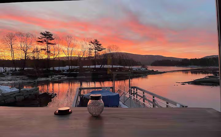 Sunset over a calm harbor with pink clouds, silhouetted trees, and a wooden deck with a table and vase in foreground
