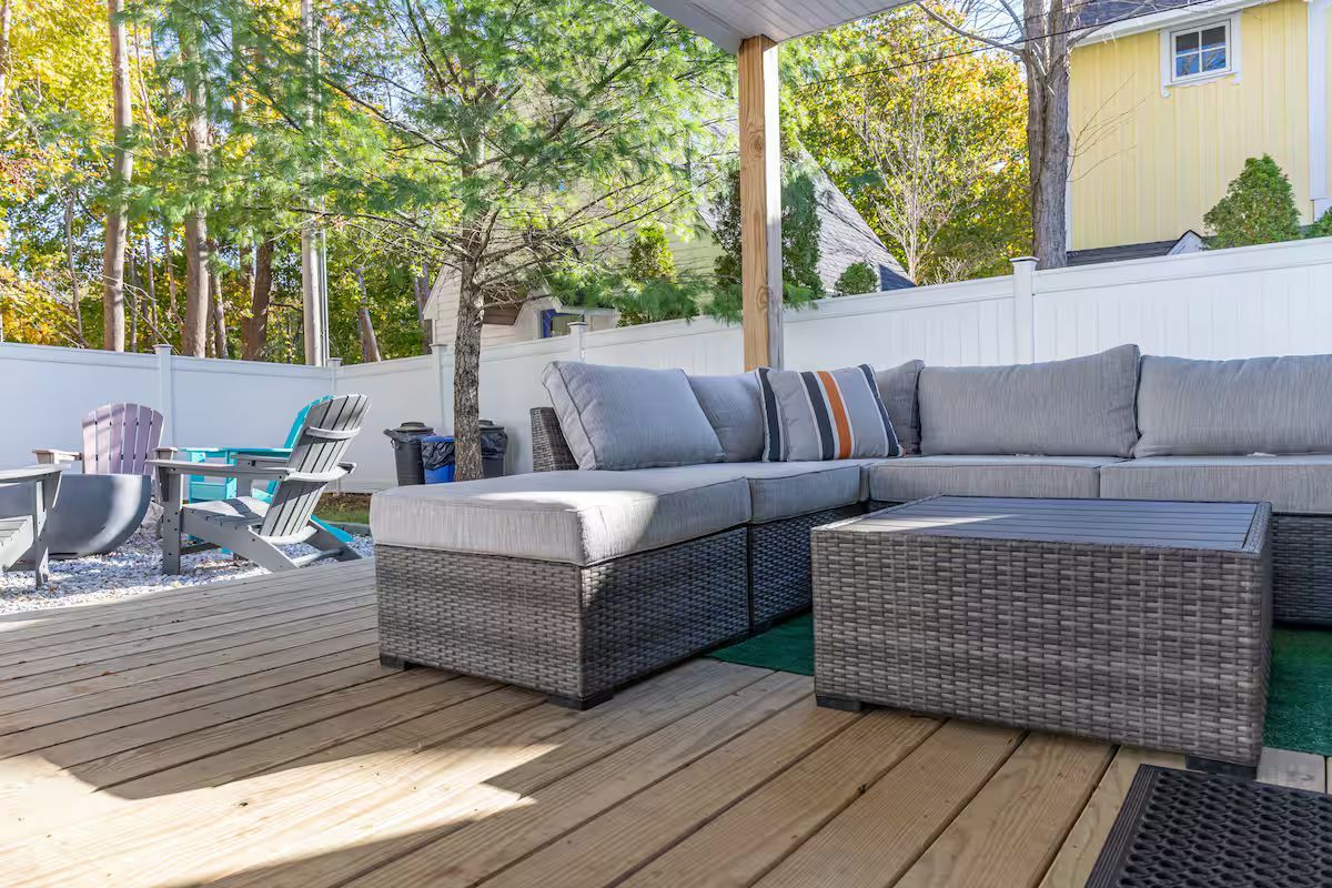 Patio with gray wicker sectional and dining table on a sunlit wooden deck under a pergola