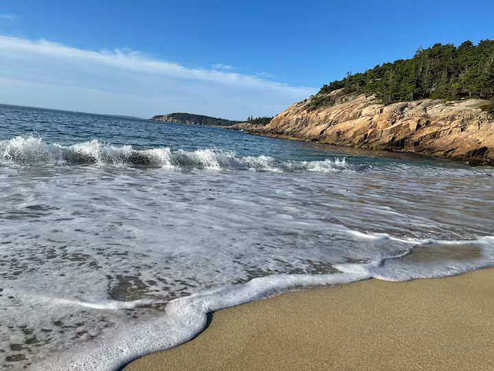 Sunny beach with waves washing onto tan sand beside a rocky, tree-lined shoreline