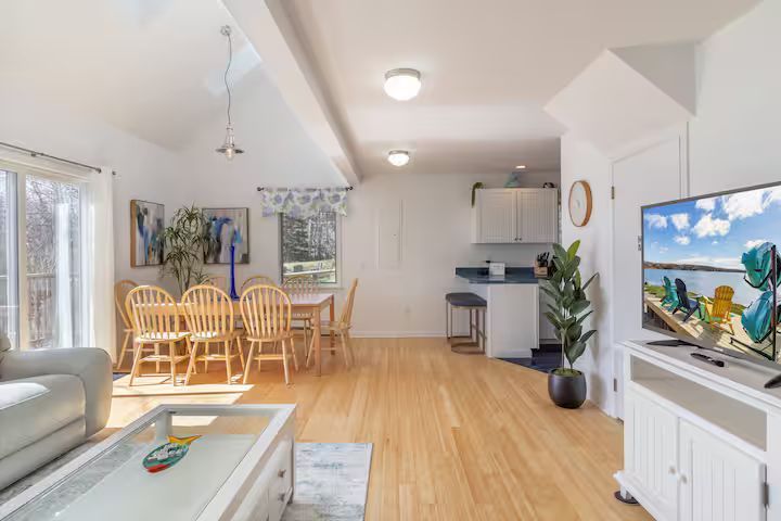 Bright open-plan living room and dining area with light wood floors, white walls, and a TV near a kitchen.