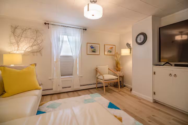 Bright living room with white sofa, yellow pillows, chair, lamp, wall art, and TV console