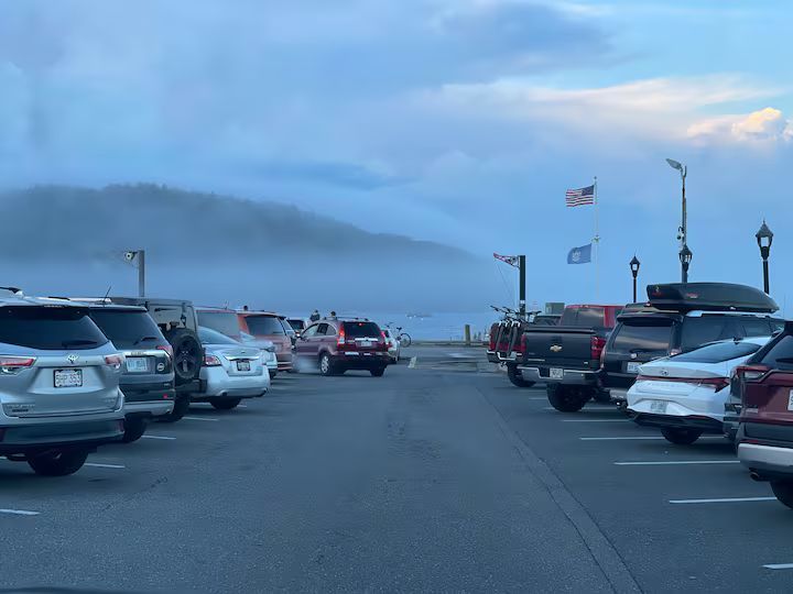 Parked cars lining a foggy waterfront road with hills in the distance.