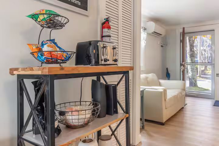Narrow hallway with a decorative table, toaster oven, fruit basket, and a bright living room beyond