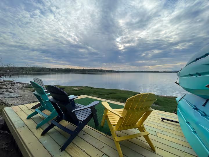 Colorful Adirondack chairs on a deck beside a calm lake under a cloudy sky