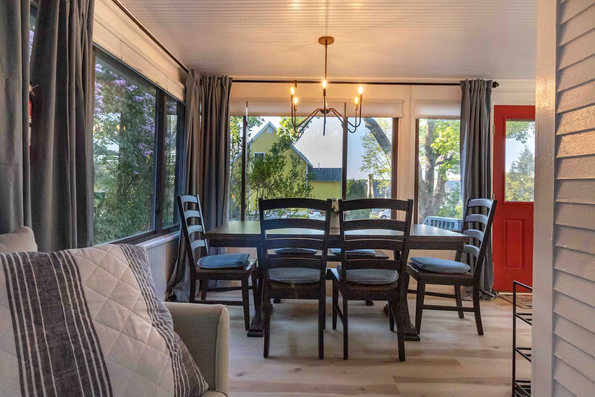 Modern dining room with a glass table, black chairs, and large windows overlooking trees and a red door