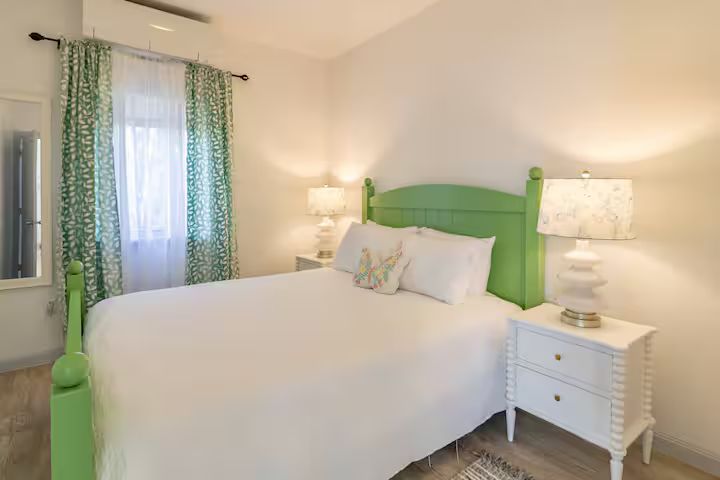 Bright bedroom with a green headboard, white bedding, and matching bedside lamps, beside a window with curtains