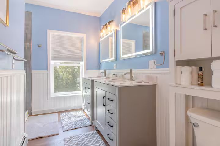Bright blue bathroom with white vanity, mirror lights, and window by a shower and toilet.
