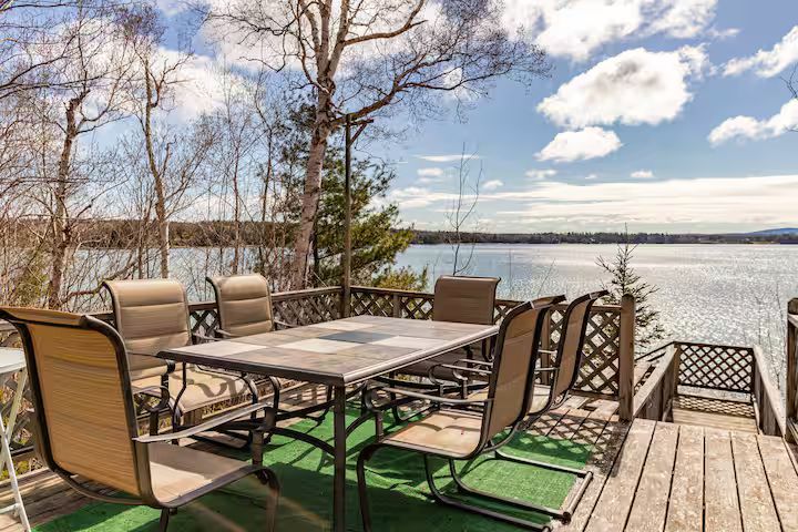 Patio dining set on a lakeside deck with chairs, table, and water view under a partly cloudy sky