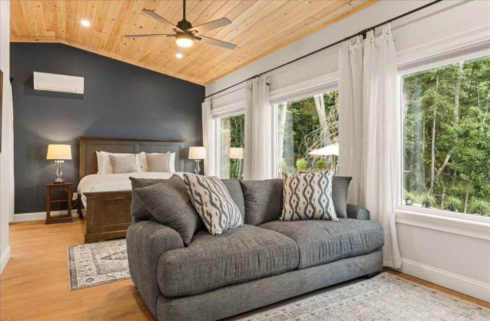 Bright bedroom with a gray sofa, wood ceiling, dark accent wall, and large windows overlooking greenery