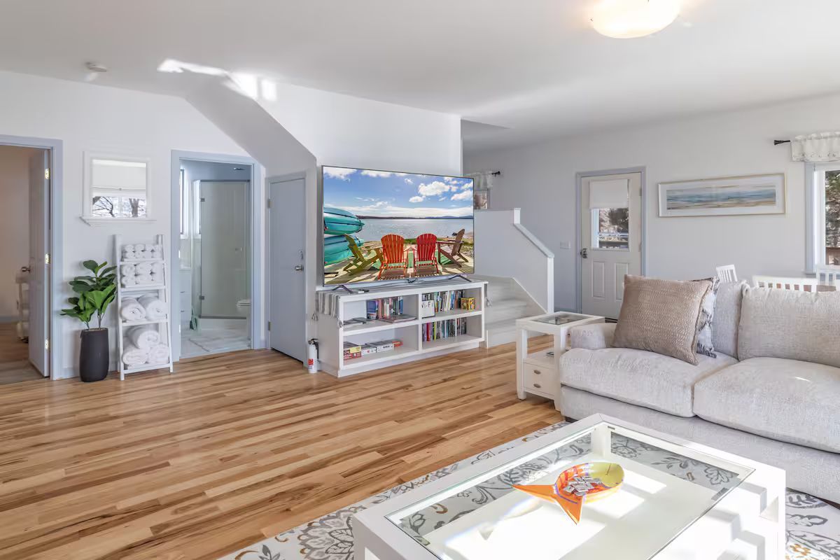 Bright living room with hardwood floors, white walls, gray sofa, white coffee table, and beach painting on TV stand