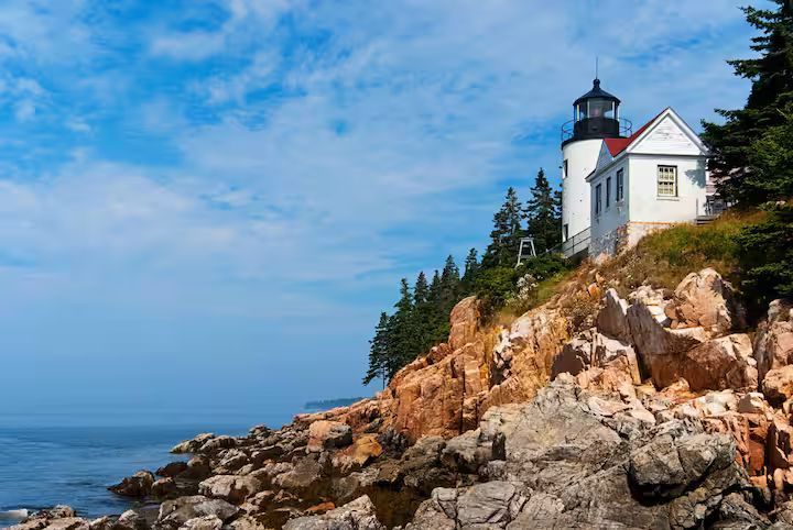 Lighthouse on a rocky coastal cliff under a blue sky with ocean at left
