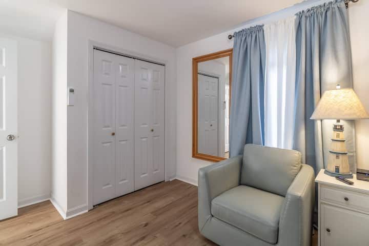 Bright bedroom corner with white closet doors, a gray chair, dresser, lamp, and blue curtains