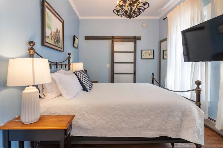 Bedroom with blue walls, white bed, wooden nightstand, chandelier, wall art, and sliding doors to a bright balcony