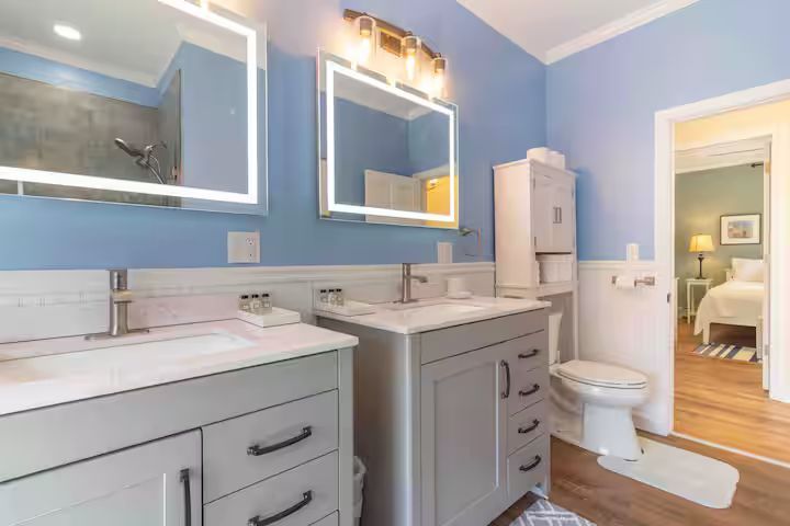 Bright blue bathroom with double sinks, mirrors, toilet, and open doorway to a bedroom