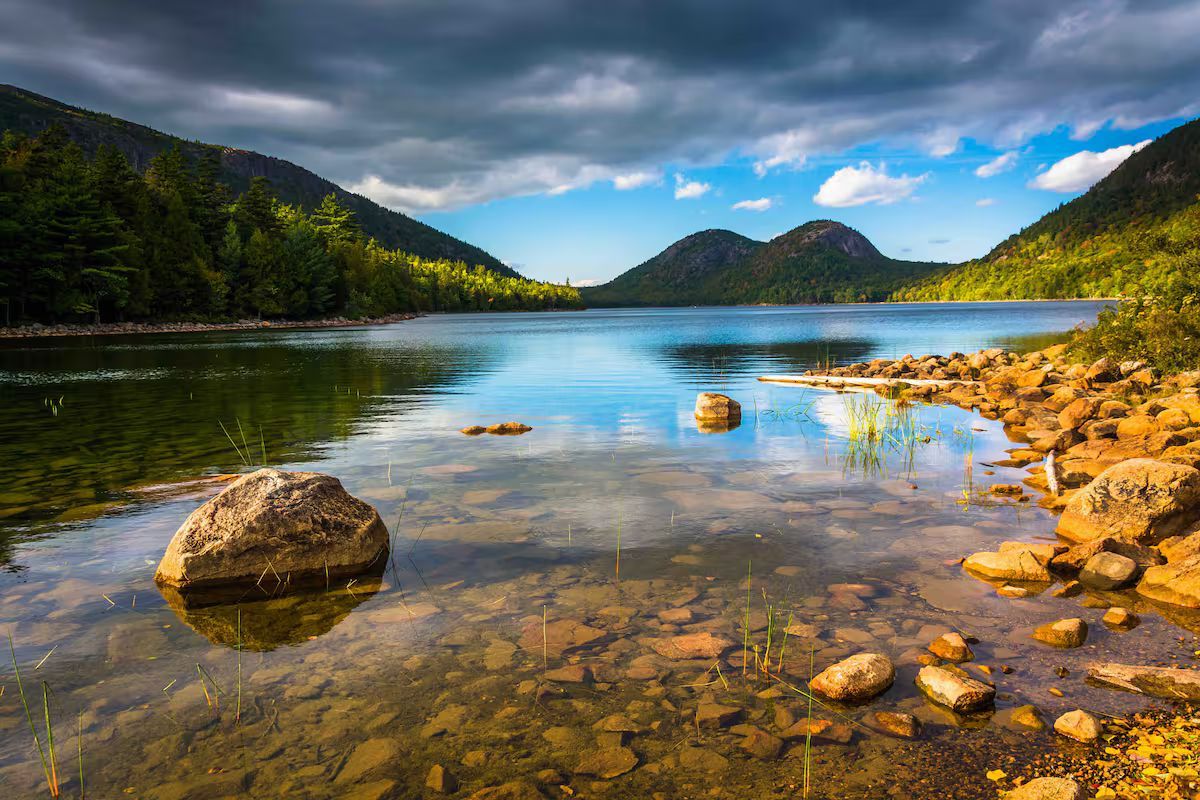 Rocky mountain lake with clear water, golden shoreline, forested hills, and blue sky with clouds
