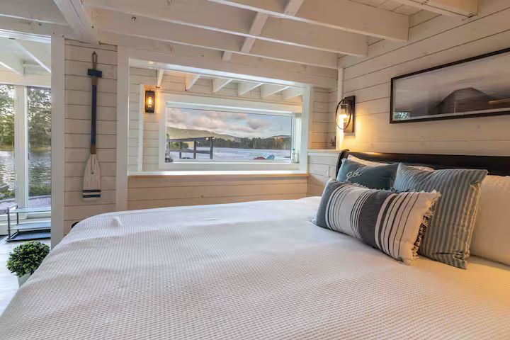 Bright bedroom with white wood walls, a large bed, and a window view of a waterfront deck