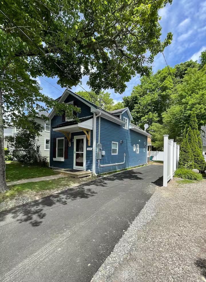 Blue house with white trim and porch, framed by trees beside a driveway under a sunny sky