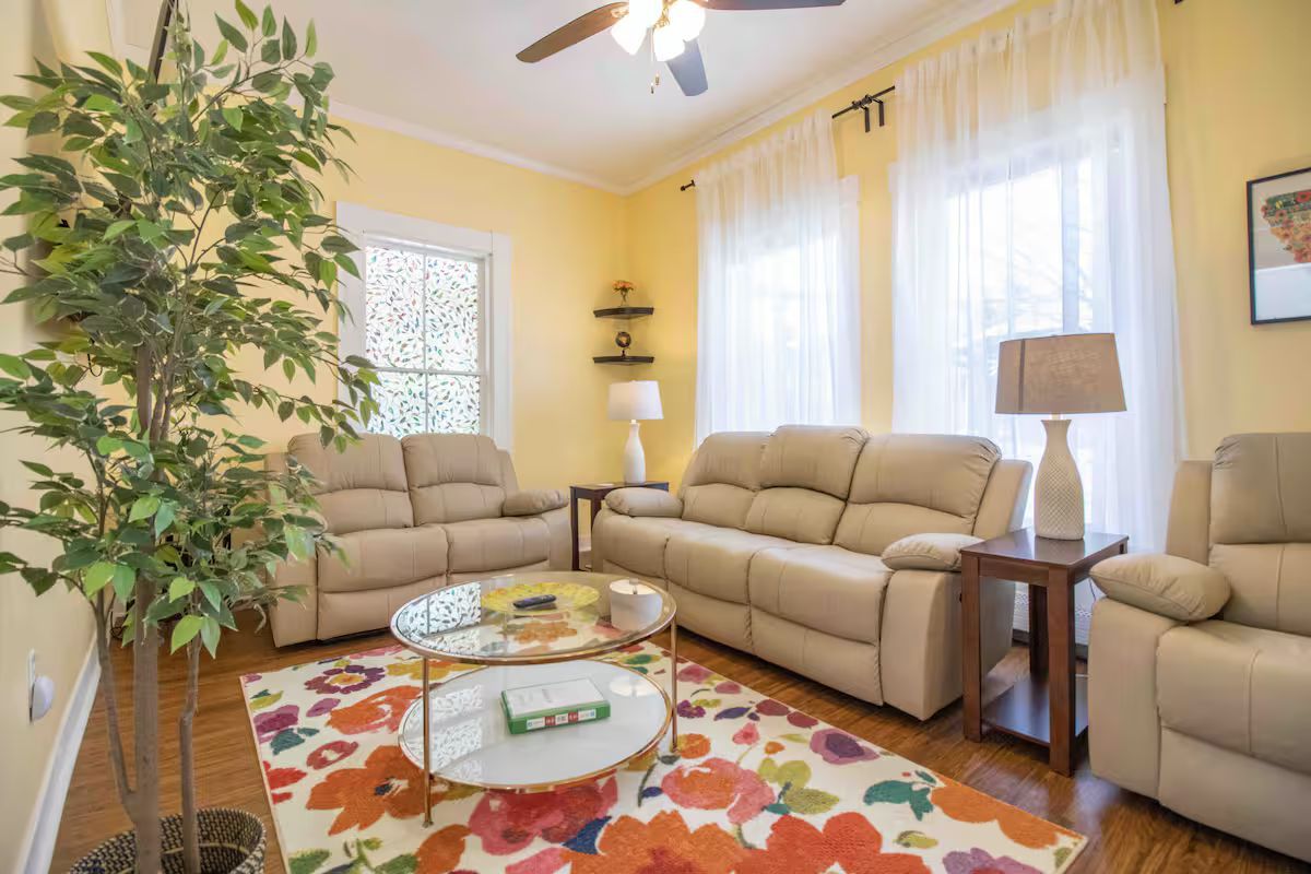 Bright living room with beige sofas, floral rug, glass coffee table, and large windows with sheer curtains