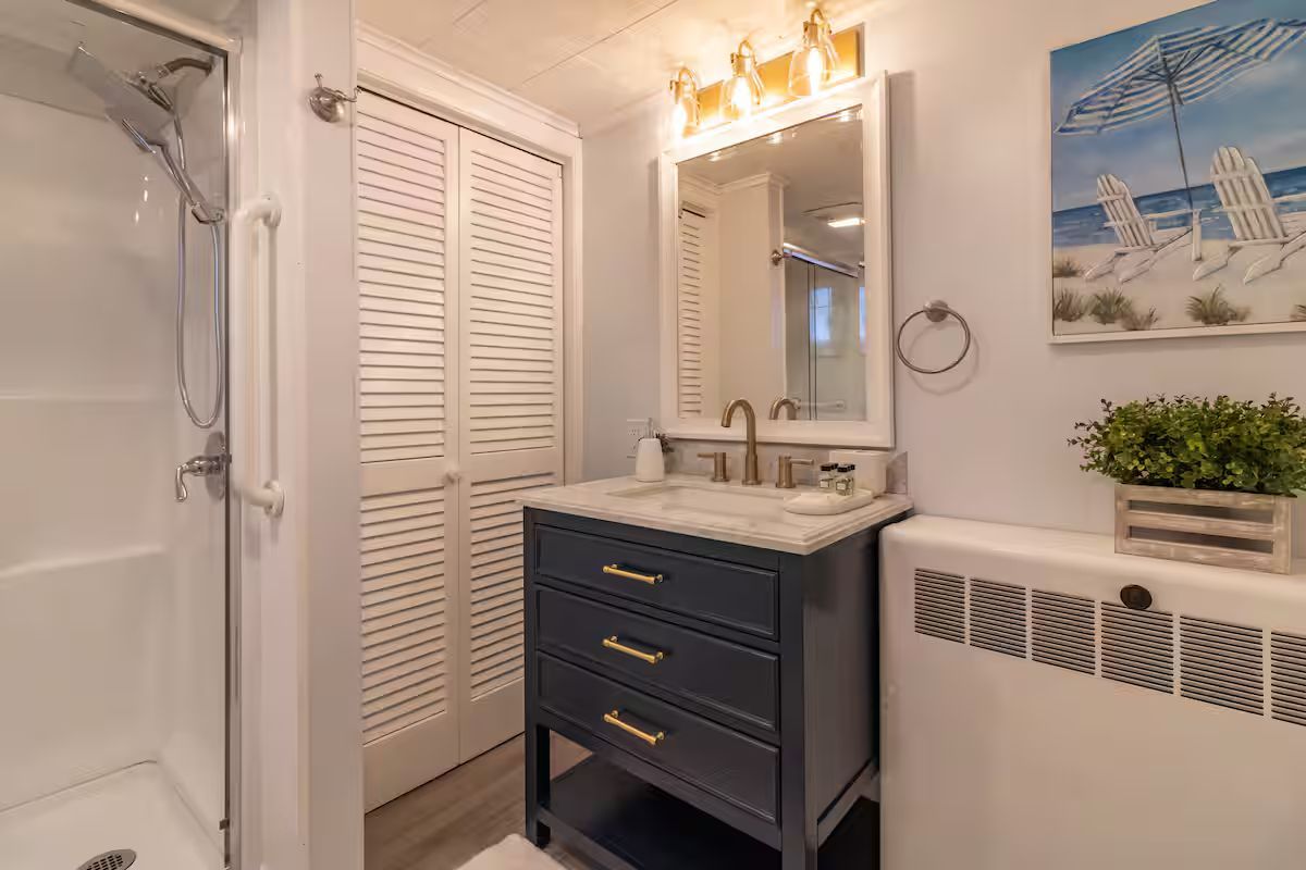 Bright bathroom vanity with navy cabinet, white shiplap doors, mirror lights, shower, and beach wall art