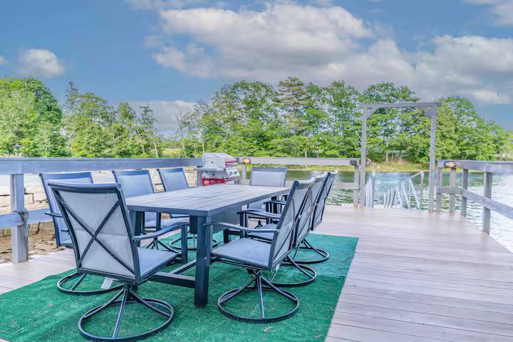Outdoor deck with patio table and chairs on a green rug, overlooking trees and water under a blue sky