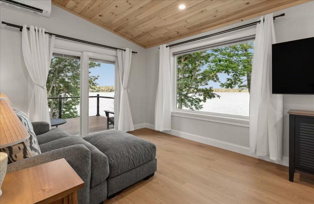 Bright living room with gray chaise, large windows, wood ceiling, and a fireplace edge