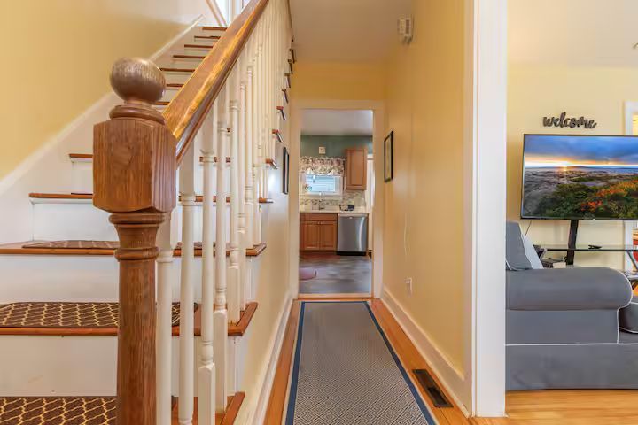 Hallway with wooden staircase, runner rug, and view into a kitchen and living room.