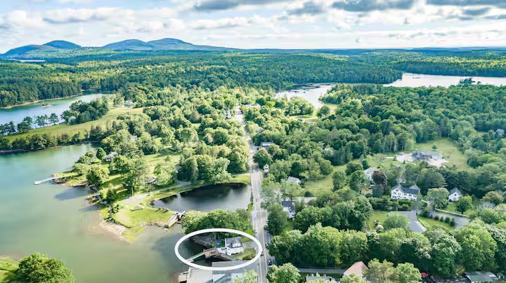 Aerial view of a green lakeside park with a bridge, winding paths, and distant mountains under a bright sky