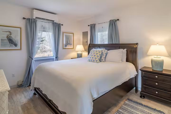 Bright bedroom with a made bed, nightstands, lamps, windows, and framed wall art
