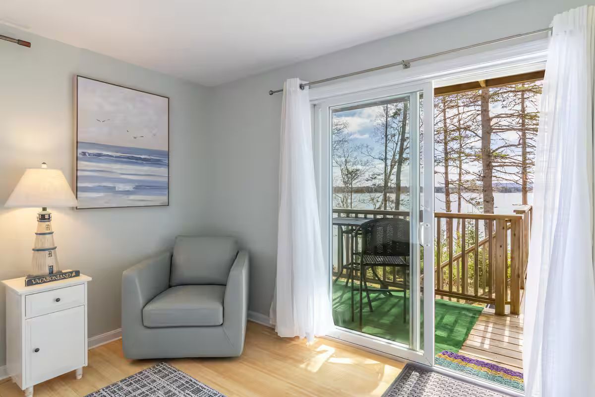 Bright living room with gray chair, lamp, artwork, and open sliding doors to a deck with trees outside