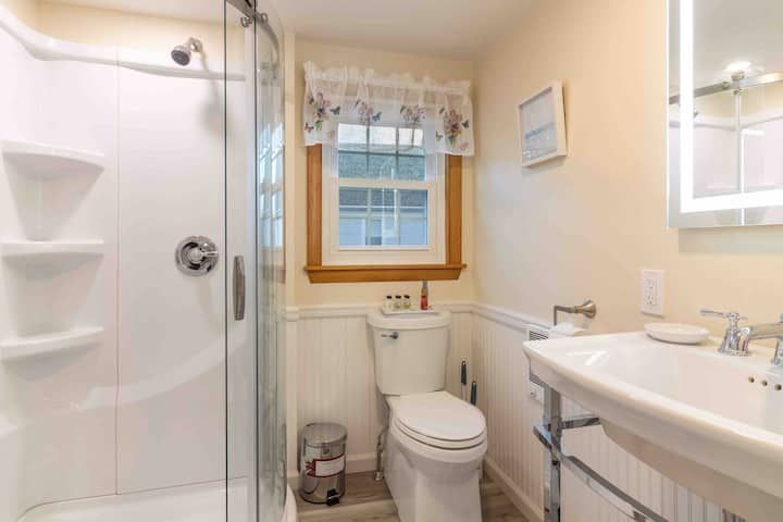 Bright small bathroom with shower, toilet, vanity sink, and window with white curtains
