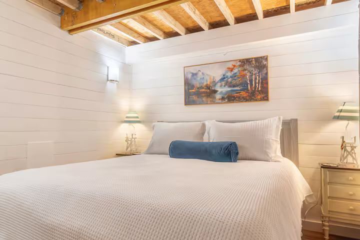 Bright bedroom with white bed, blue bolster, wall art, bedside lamps, and exposed wooden ceiling beams