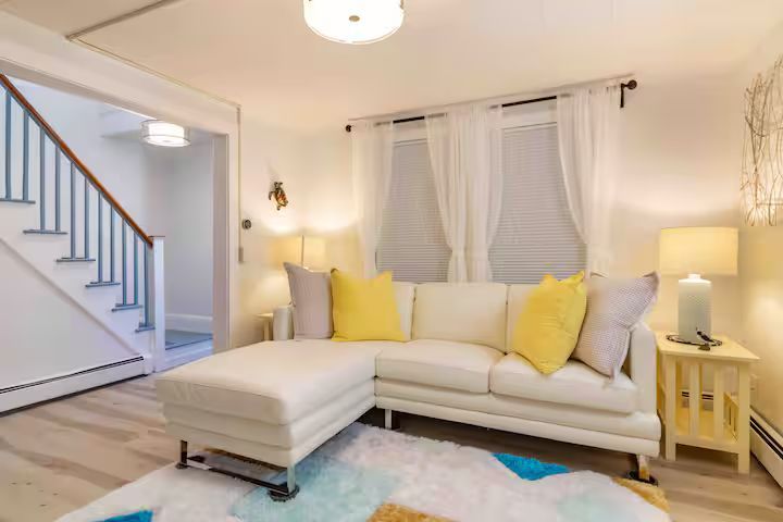 Bright living room with white sofa, yellow pillows, ottoman, lamp, staircase, and large window blinds