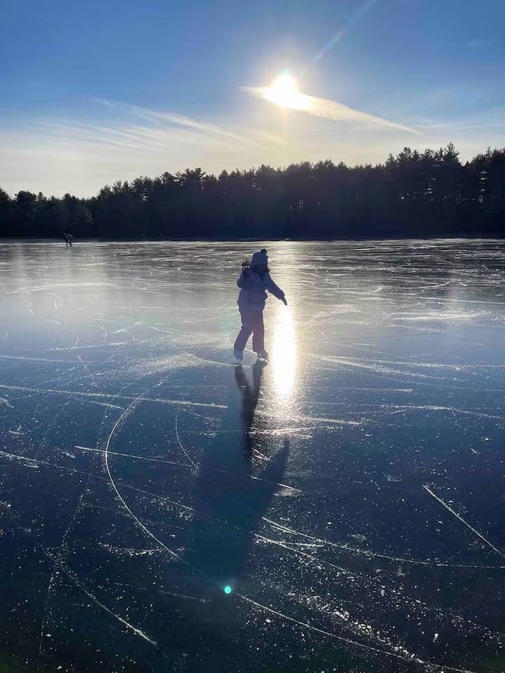 Person ice skating on a frozen lake at sunset, with trees silhouetted in the background.