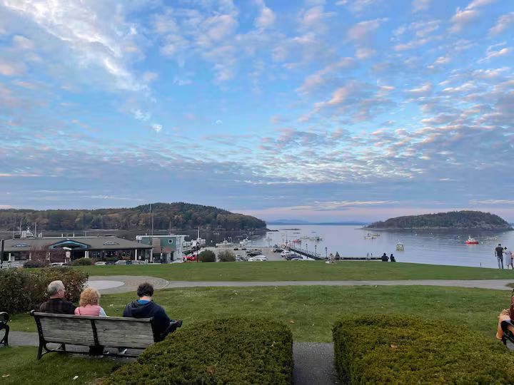 People sitting on a bench overlooking a calm harbor with small islands under a cloudy sky.