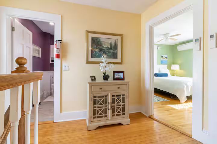 Bright hallway with wood floors, framed art, and open doors to a purple room and a sunlit bedroom