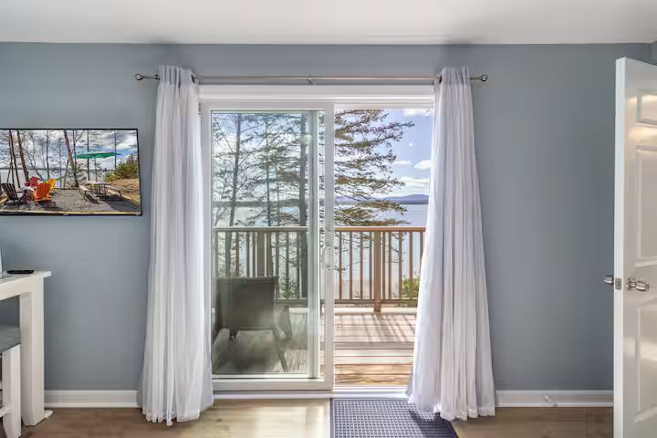 Blue bedroom with white curtains framing sliding glass doors to a balcony and trees outside