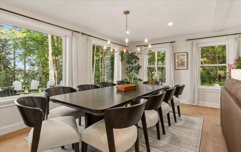 Bright dining room with a large dark table, black chairs, and floor-to-ceiling windows overlooking greenery