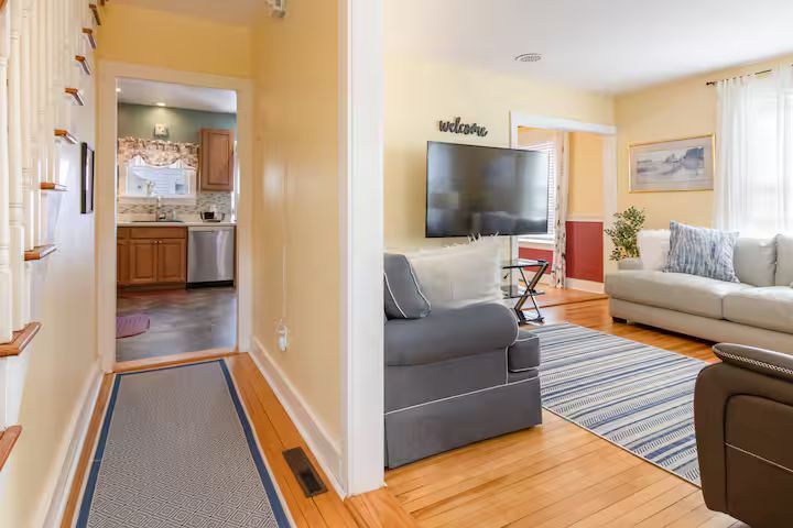 Bright living room with gray and beige sofas, TV, striped rug, and a view into a kitchen hallway.