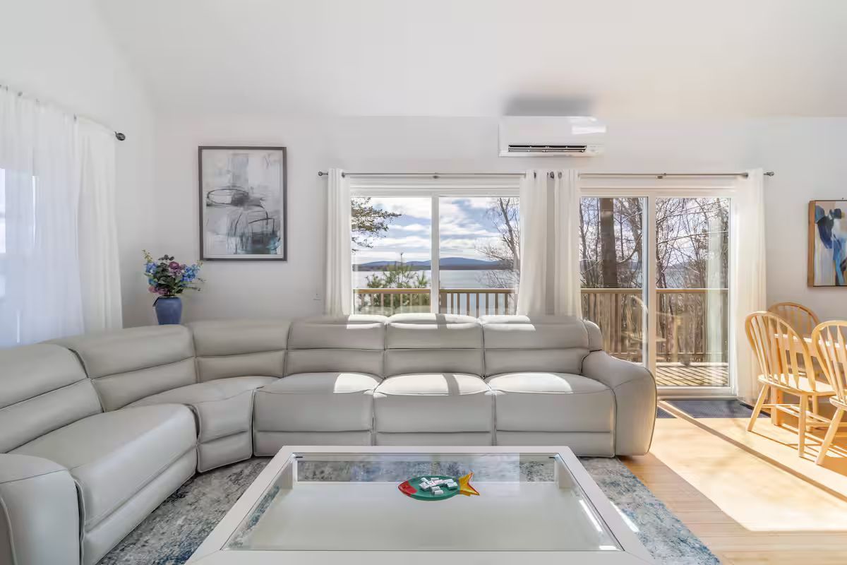 Bright living room with sectional sofa, glass coffee table, and open doors to a sunlit patio and waterfront view
