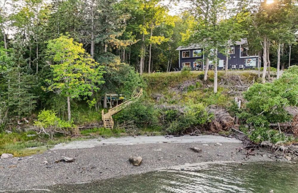 Rocky shoreline with calm water, wooded hillside, and a dark cabin nestled among trees.