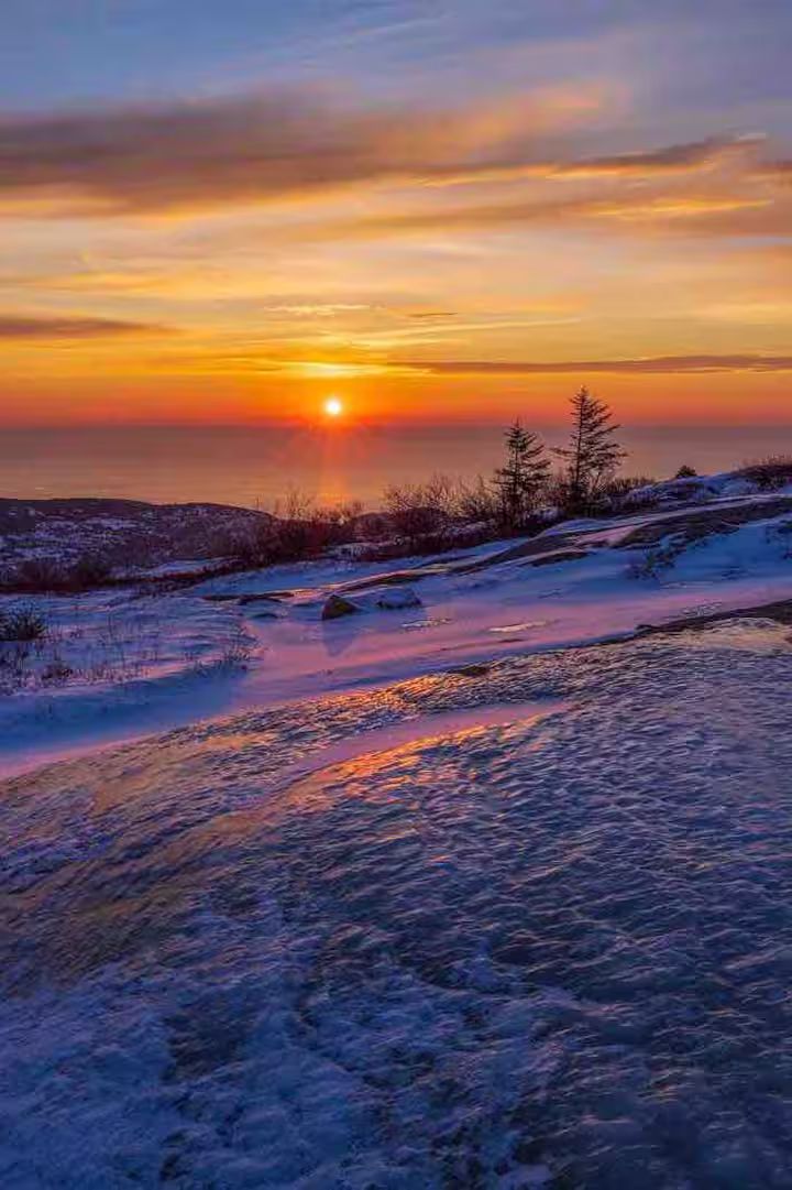 Snowy shoreline at sunset with pink-orange sky reflecting on icy water