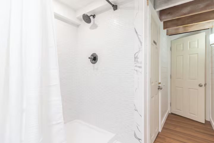 White tiled bathroom shower with curtain, black fixtures, and an open door to a hall with wood floors.