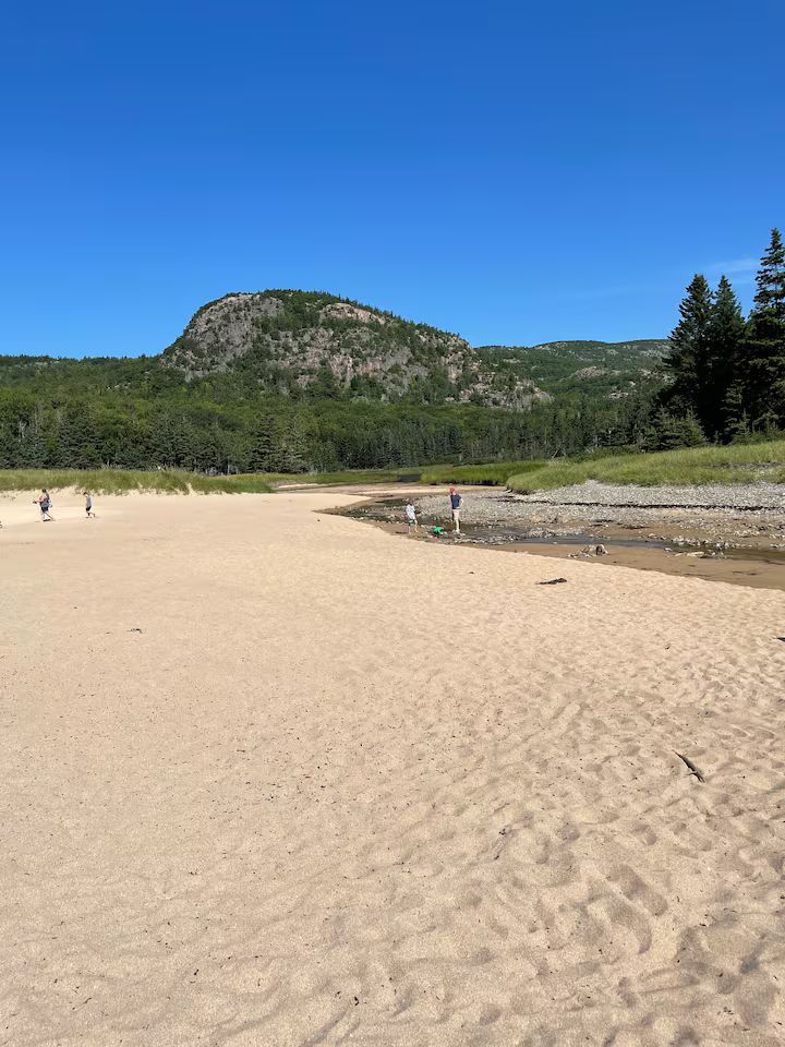 Wide sandy beach with a few people, forest, and a rocky hill under a clear blue sky