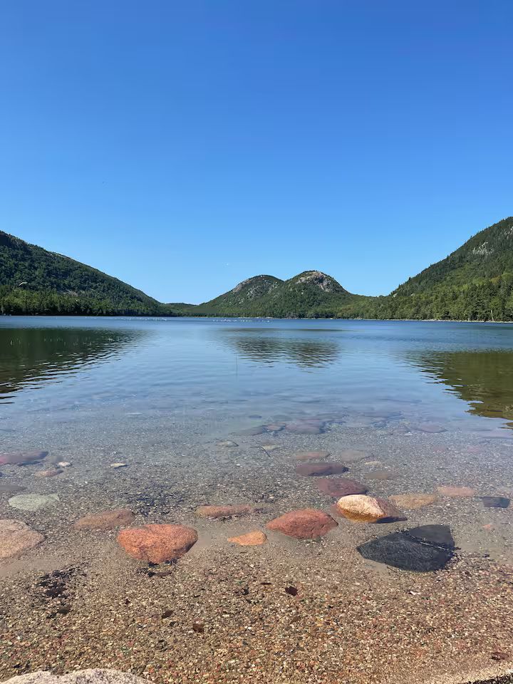 Clear lake with pebbled shore, calm reflective water, and green hills under a bright blue sky.