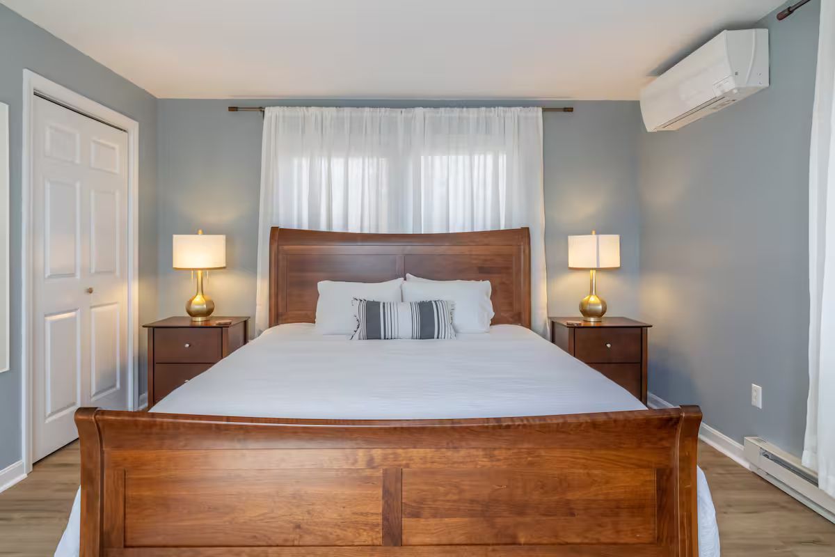 Cozy bedroom with wooden bed, white bedding, bedside lamps, and a window with sheer curtains.