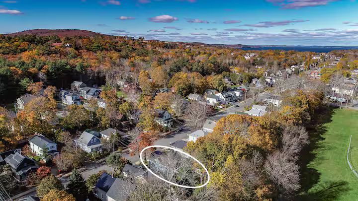 Aerial view of a suburban neighborhood with autumn trees and a circled roof near a green field.