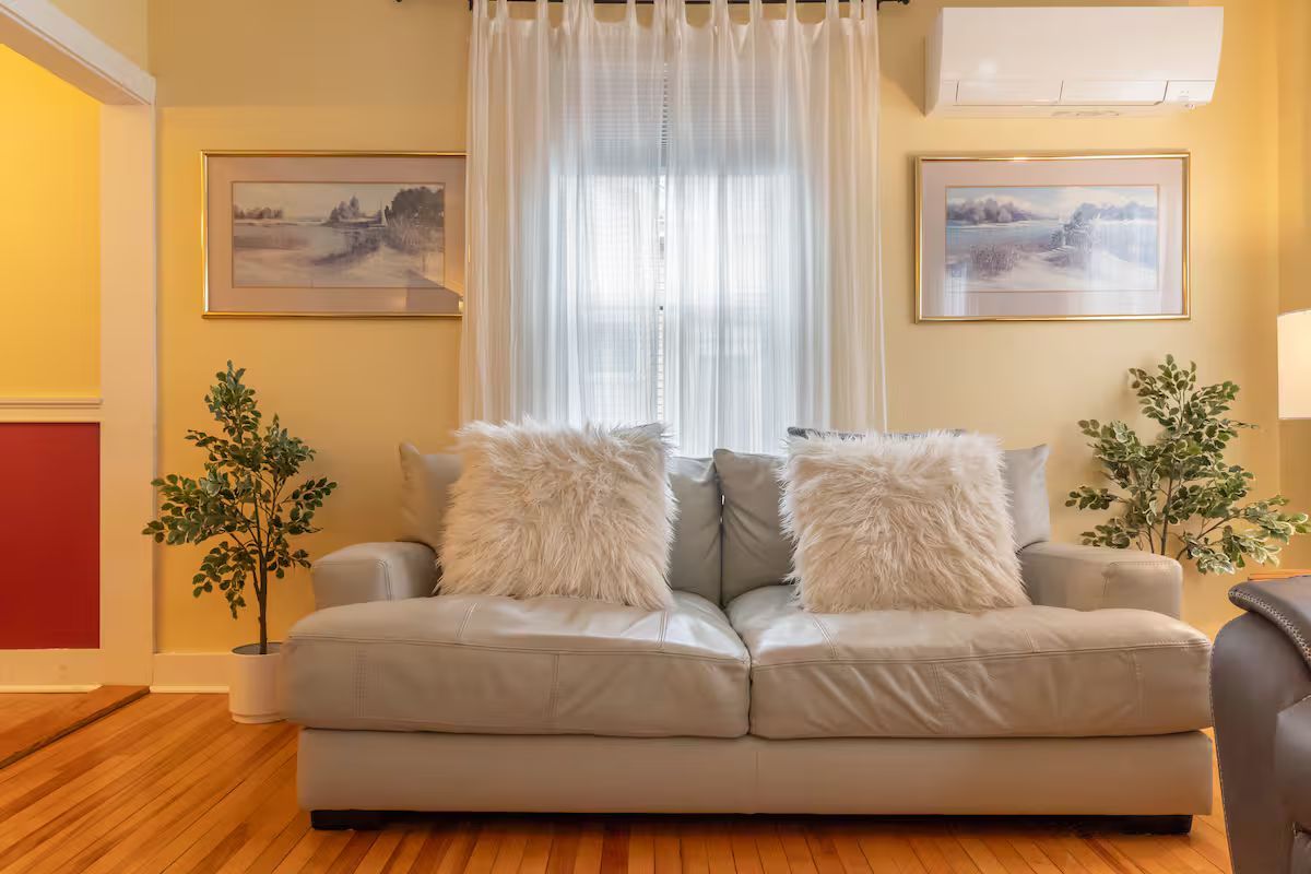 Bright living room with beige sofa, fluffy pillows, framed art, plants, and a sheer-curtained window