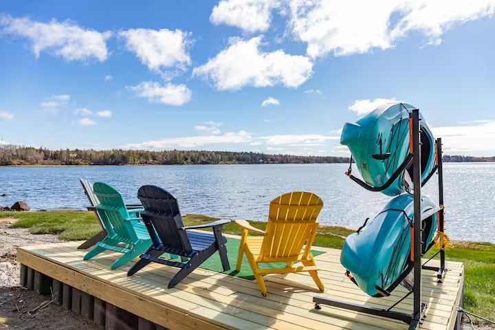 Colorful Adirondack chairs on a dock by a calm lake under a blue sky, with kayaks stacked nearby.